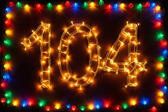 Warme lichtjes vormen 104 op een donkere muur, omkaderd door kleurrijke feestlichtjes met onscherpe lichtvlekken.