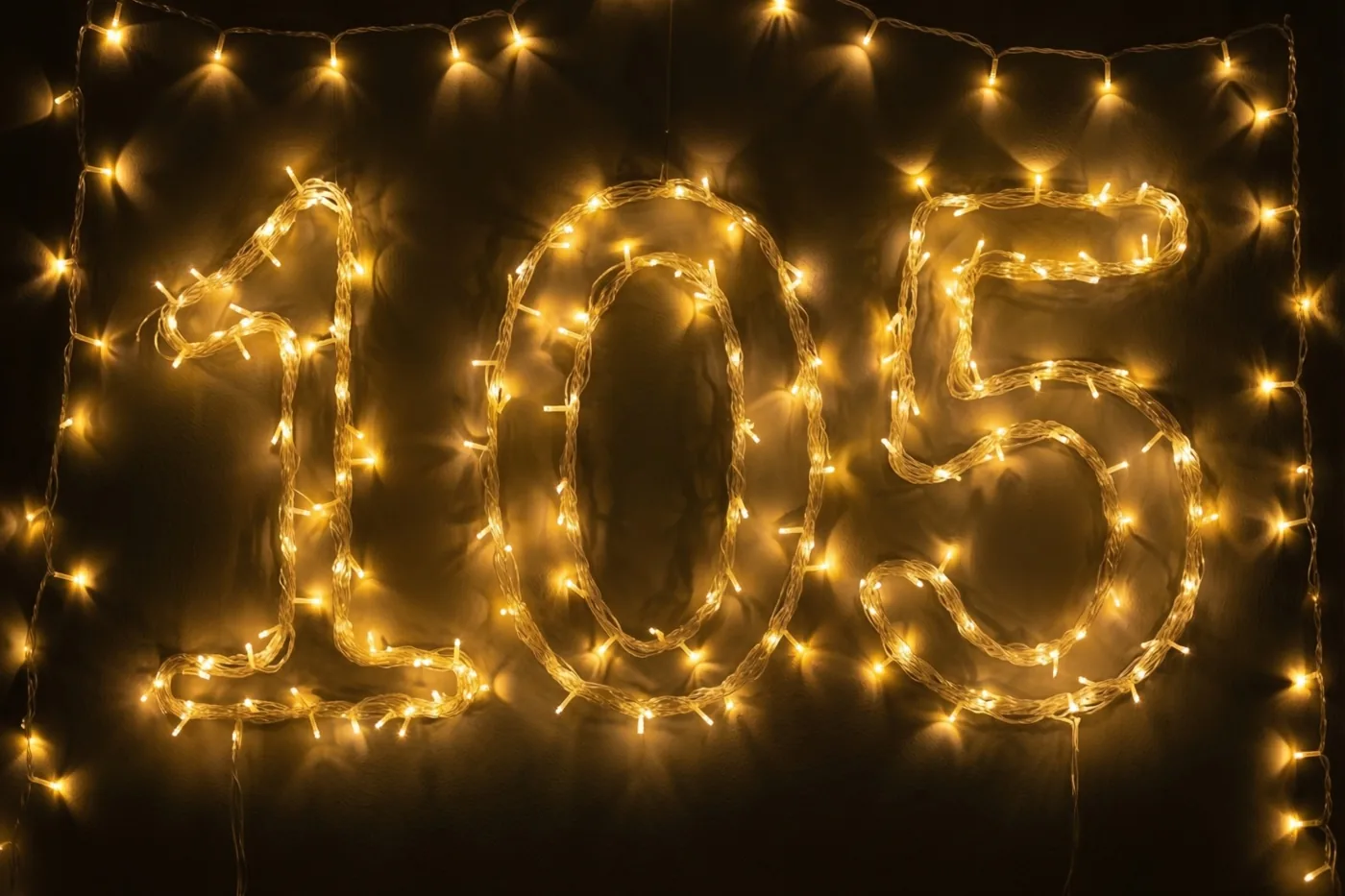 Warme lichtjes vormen 105 tegen een donkere muur, met goudgele onscherpe lichtvlekken eromheen.