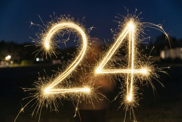 Gouden lichtsporen van sterretjes vormen 24 tegen een donkere nachthemel, met onscherpe lichtjes op de achtergrond.