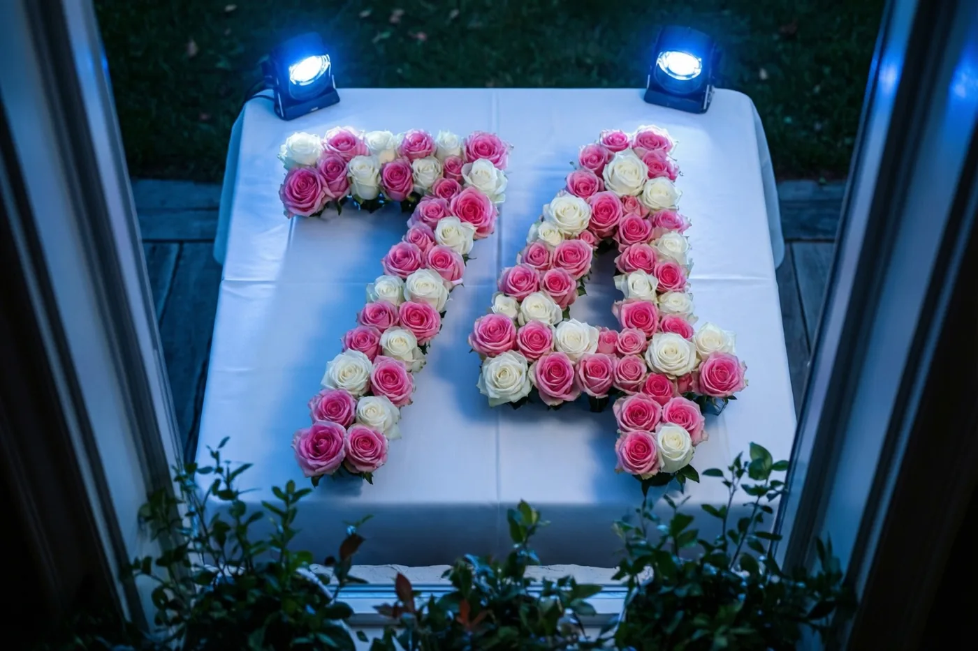 Van bovenaf is 74 van roze en witte rozen te zien op een wit tafelkleed onder blauwe spots, omkaderd door raamruiten met groen onderaan.