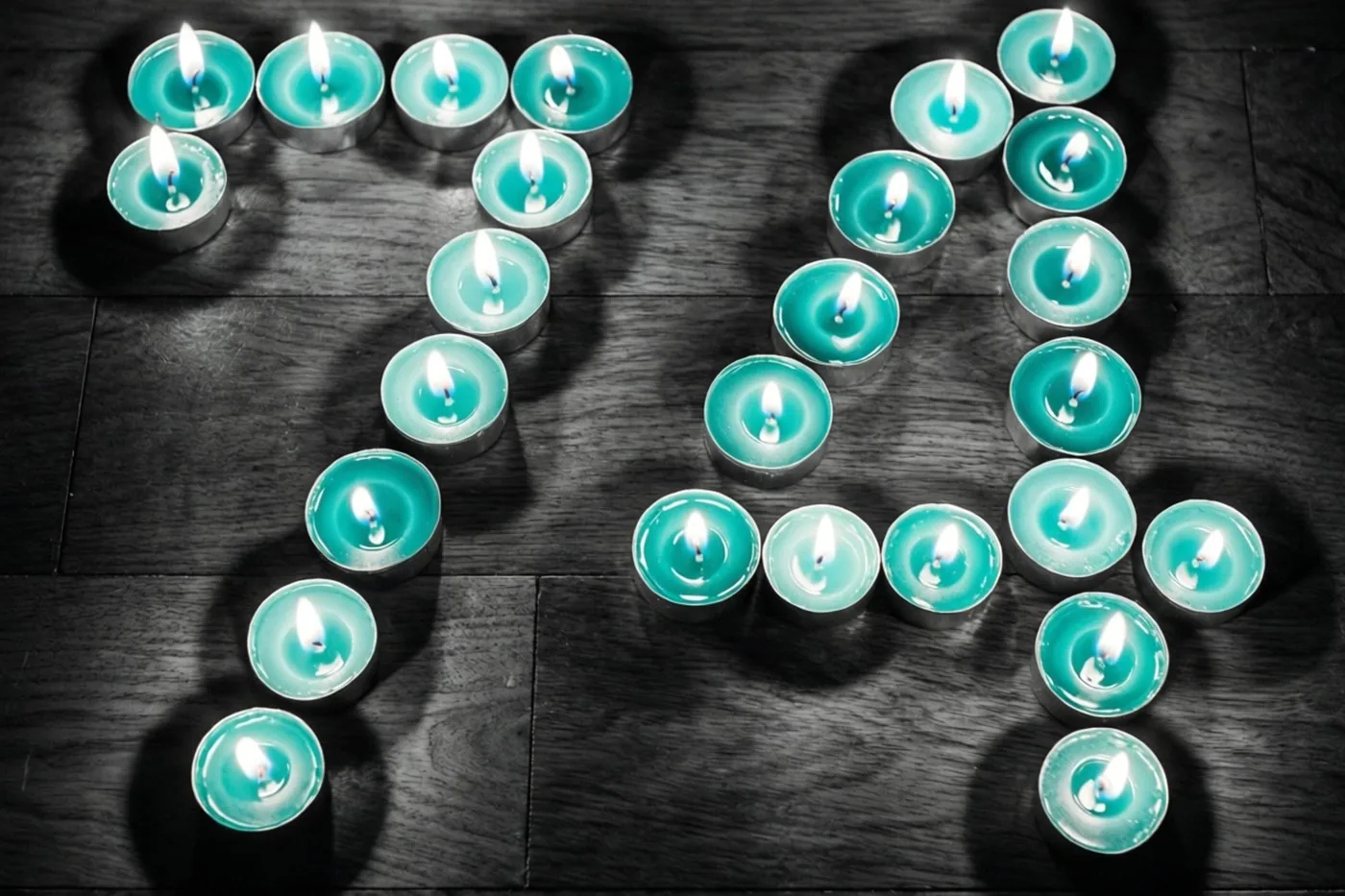 Brandende theelichtjes vormen 74 op een donkere houten vloer in een sober beeld met blauwgroene en zwarte tinten.