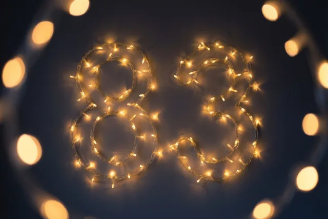 Warme lichtjes vormen het getal 83 op een donkere muur, met onscherpe feestlichtjes op de achtergrond.