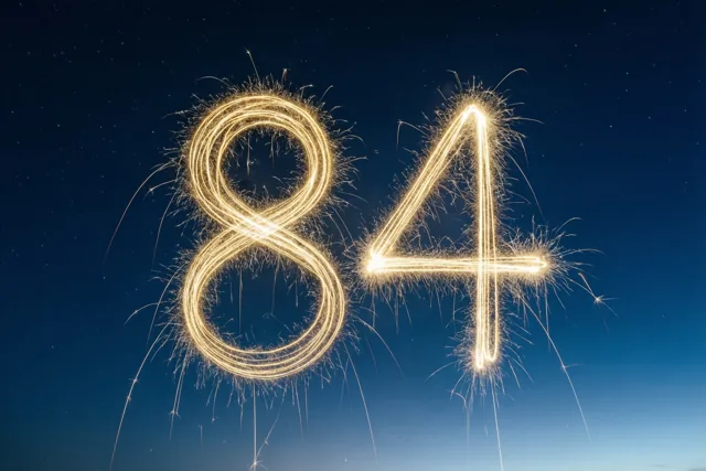 Gouden vonkensporen vormen het getal 84 tegen een donkere hemel met zichtbare sterren.