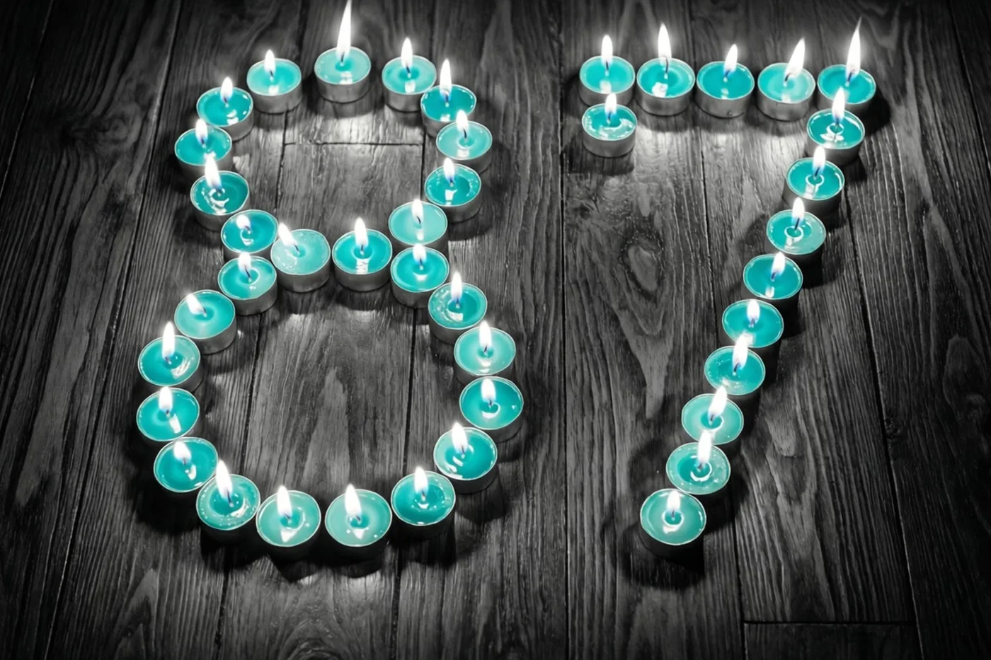 Theelichtjes vormen het getal 87 op een donkere houten vloer in een beeld met blauwgroene en zwarte tinten.