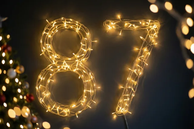 Warme lichtjes vormen het getal 87 tegen een donkere muur, met onscherpe lichtvlekken en links een wazige kerstboom.