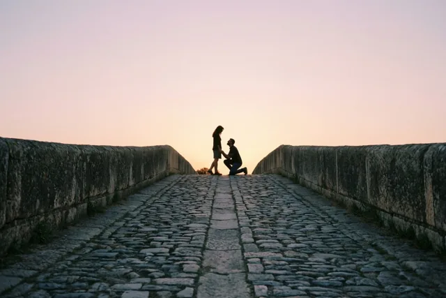 Een man knielt voor zijn partner op een brug met kasseien, met hun silhouetten tegen een roze en oranje avondlucht.