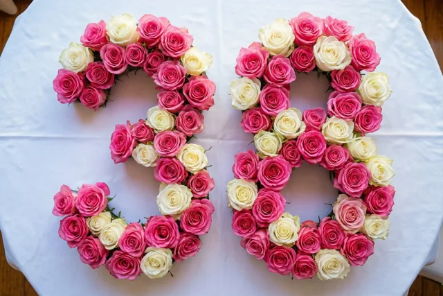 Van bovenaf zie je het getal 38 gevormd door roze en witte rozen op een wit tafelkleed.