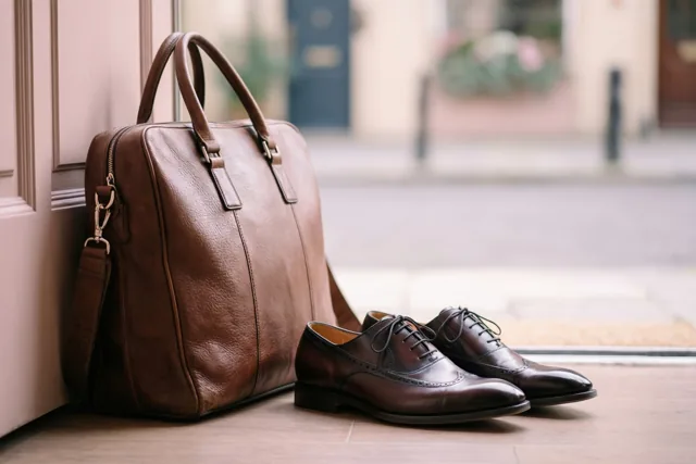 Een bruine leren aktetas en een paar gepoetste bruine geklede schoenen staan buiten op een tegelvloer naast een deur, met een onscherpe straat op de achtergrond.
