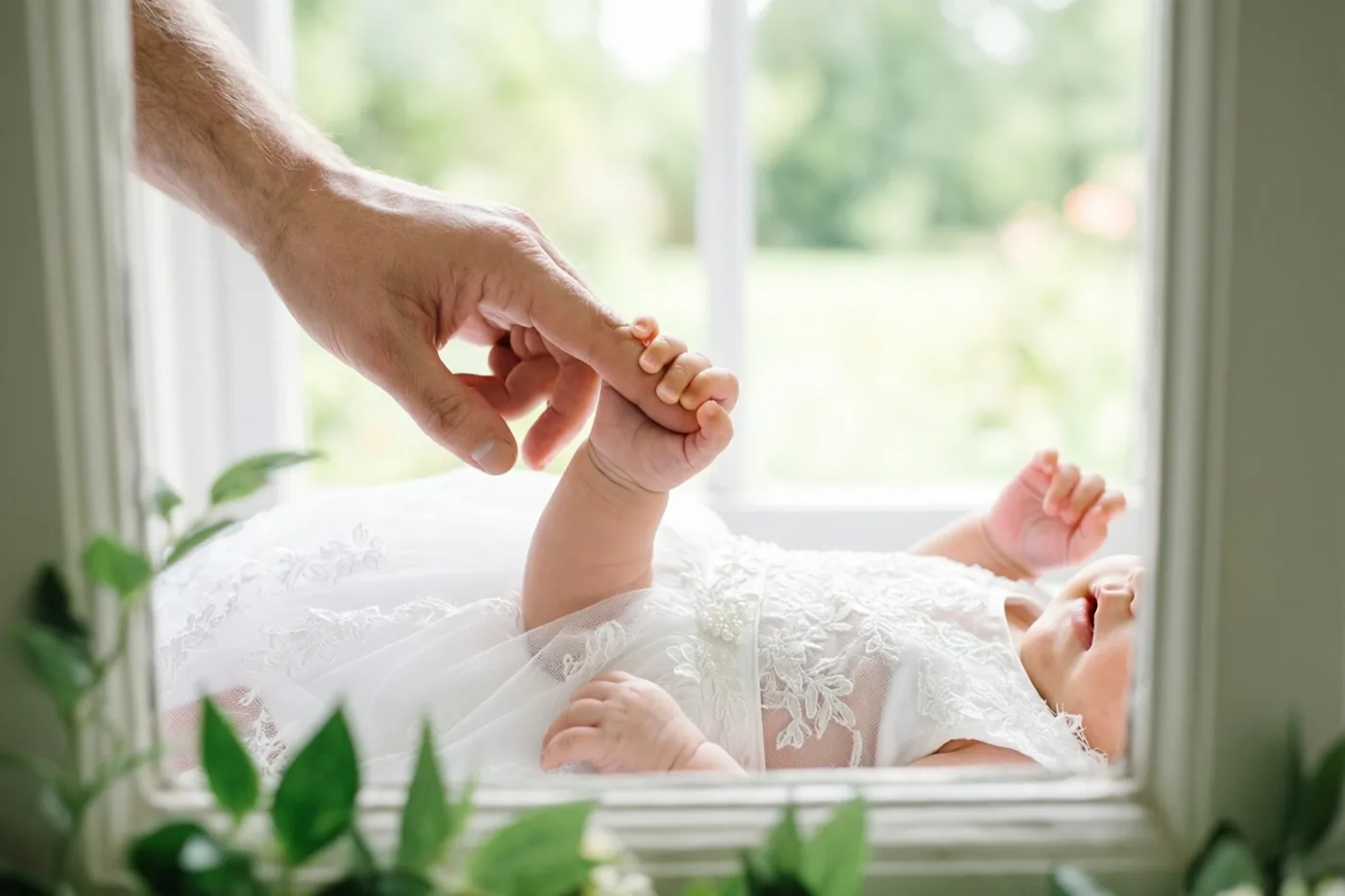 Een baby in een wit kanten doopkleed ligt op een vensterbank en houdt een vinger van een volwassene vast, bij zacht daglicht met groene bladeren op de voorgrond.