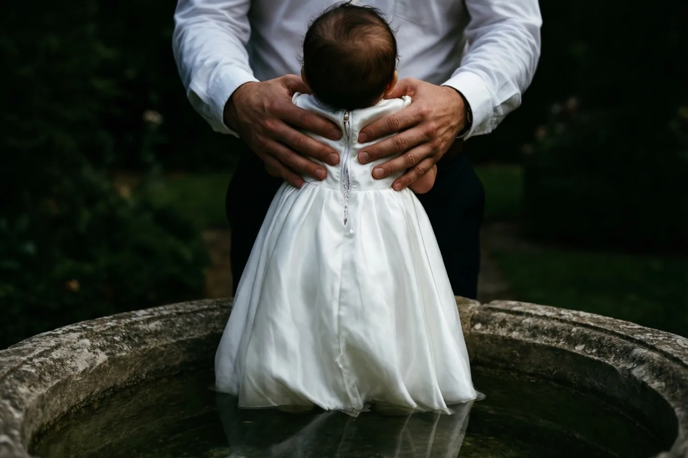 Een ouder in een wit hemd houdt een baby in een lang wit kleed boven de rand van een stenen doopvont met water, met donkergroen gebladerte op de achtergrond.