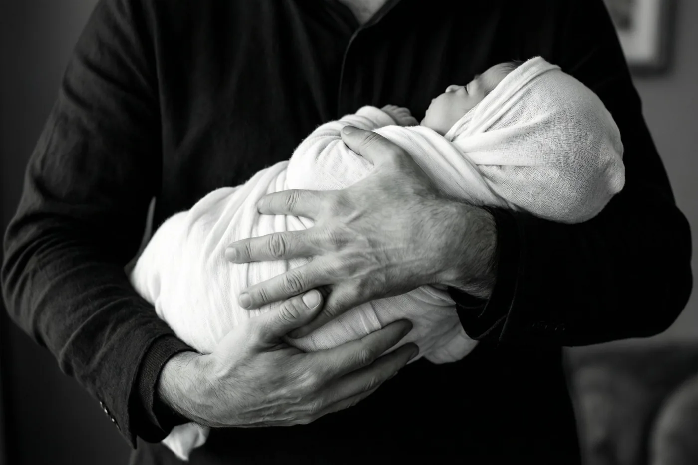 Op een zwart-witfoto houden de handen van een meter of peter een baby in een witte sjaal vast, zonder zichtbare gezichten.