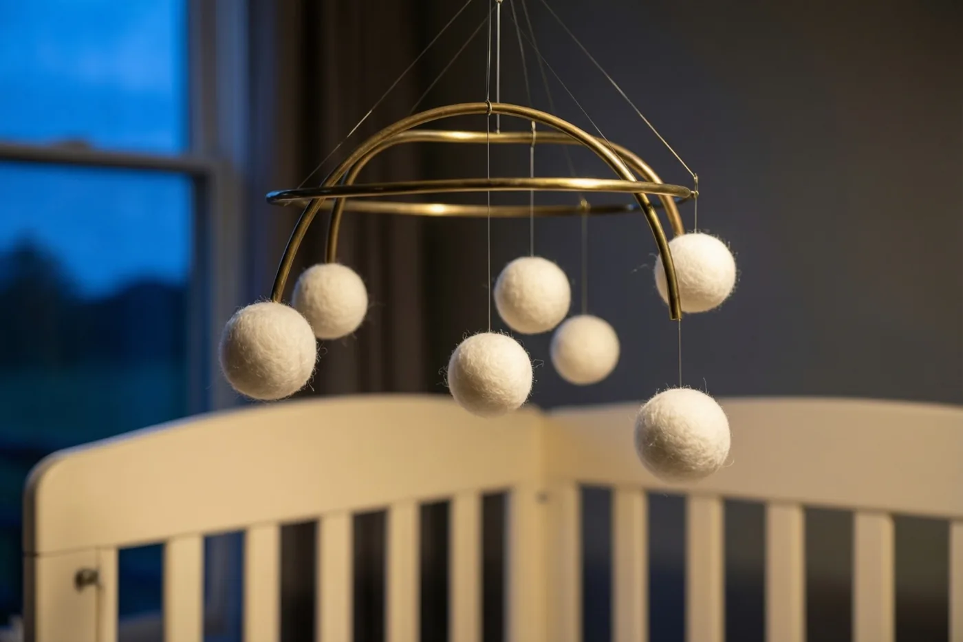 Een mobiel met messing ringen en witte vilten balletjes hangt boven een ivoorkleurig babybedje, met buiten het raam de schemering.