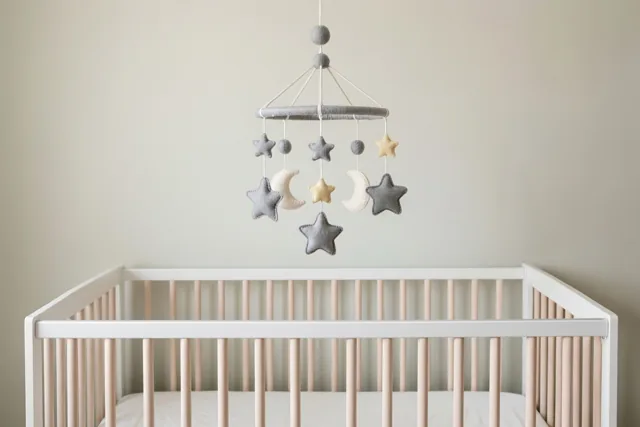 Boven een wit babybedje met lichte houten spijlen hangt een mobiel van vilten sterren en manen tegen een effen lichte muur.