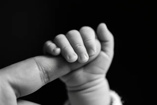 Op een zwart-witfoto grijpt het kleine handje van een pasgeboren baby de vinger van een volwassene vast tegen een zwarte achtergrond.