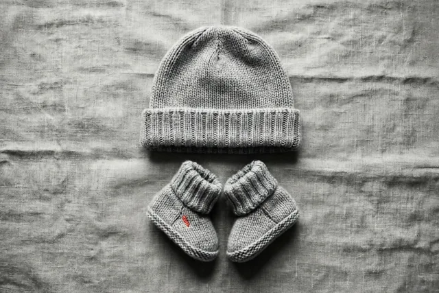 Een gebreid babymutsje en bijpassende slofjes liggen op een linnen doek met structuur, recht van bovenaf gefotografeerd in grijstinten.