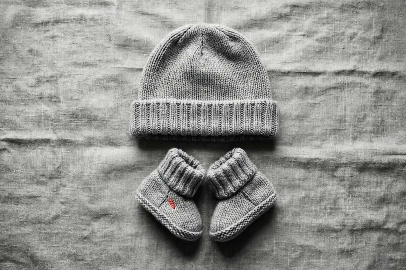 Een gebreid babymutsje en bijpassende slofjes liggen op een linnen doek met structuur, recht van bovenaf gefotografeerd in grijstinten.