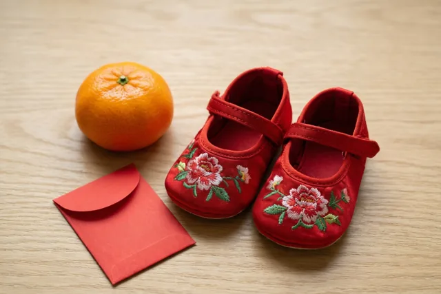 Een paar kleine rode zijden babyschoentjes met geborduurde bloemen staat op een lichte houten ondergrond naast een mandarijn en een klein rood envelopje.