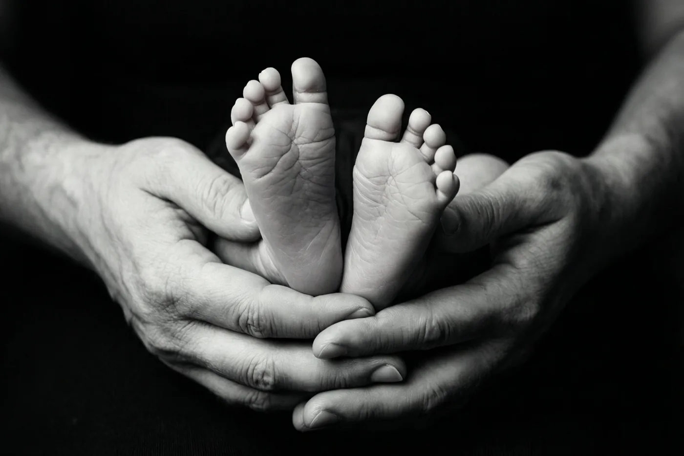 Op een zwart-witfoto ondersteunen volwassen handen de kleine voetjes van een pasgeboren baby tegen een donkere achtergrond.