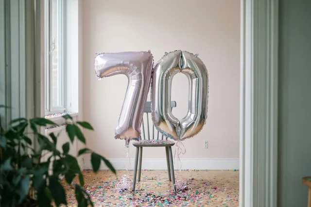 Folieballonnen in de vorm van 70 zijn vastgemaakt aan een stoel in een lichte kamer met gekleurde confetti op de vloer en links een raam.