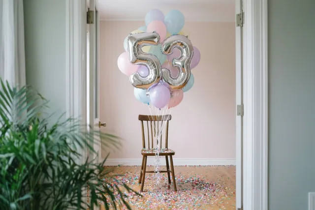 Een houten stoel met zilveren en pastelkleurige ballonnen in de vorm van 53 staat in een lichte kamer met confetti op de houten vloer, gezien door een deuropening.