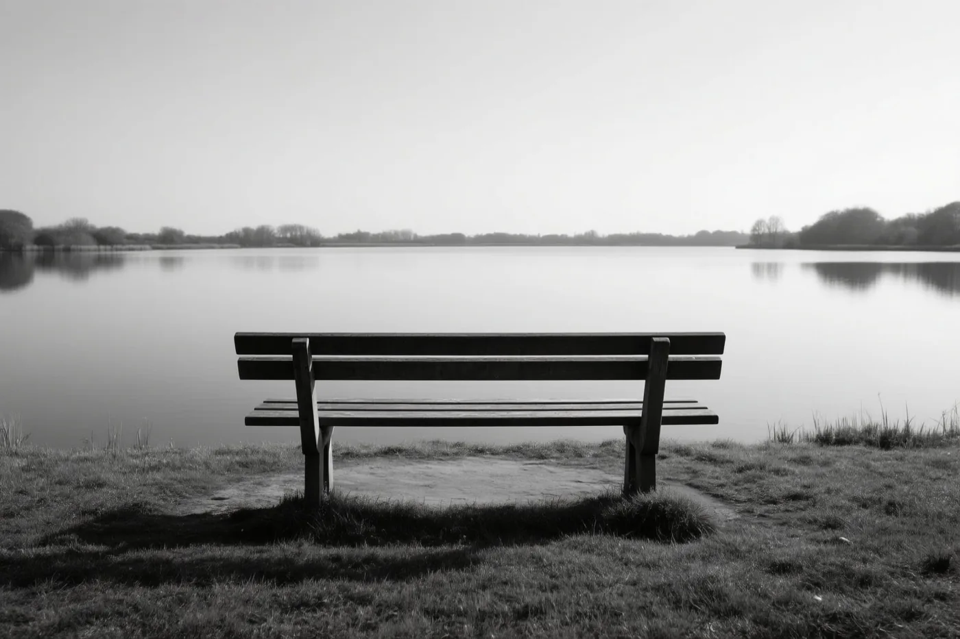 Een houten bank staat op een grasoever met uitzicht op een stil meer in een monochroom beeld.