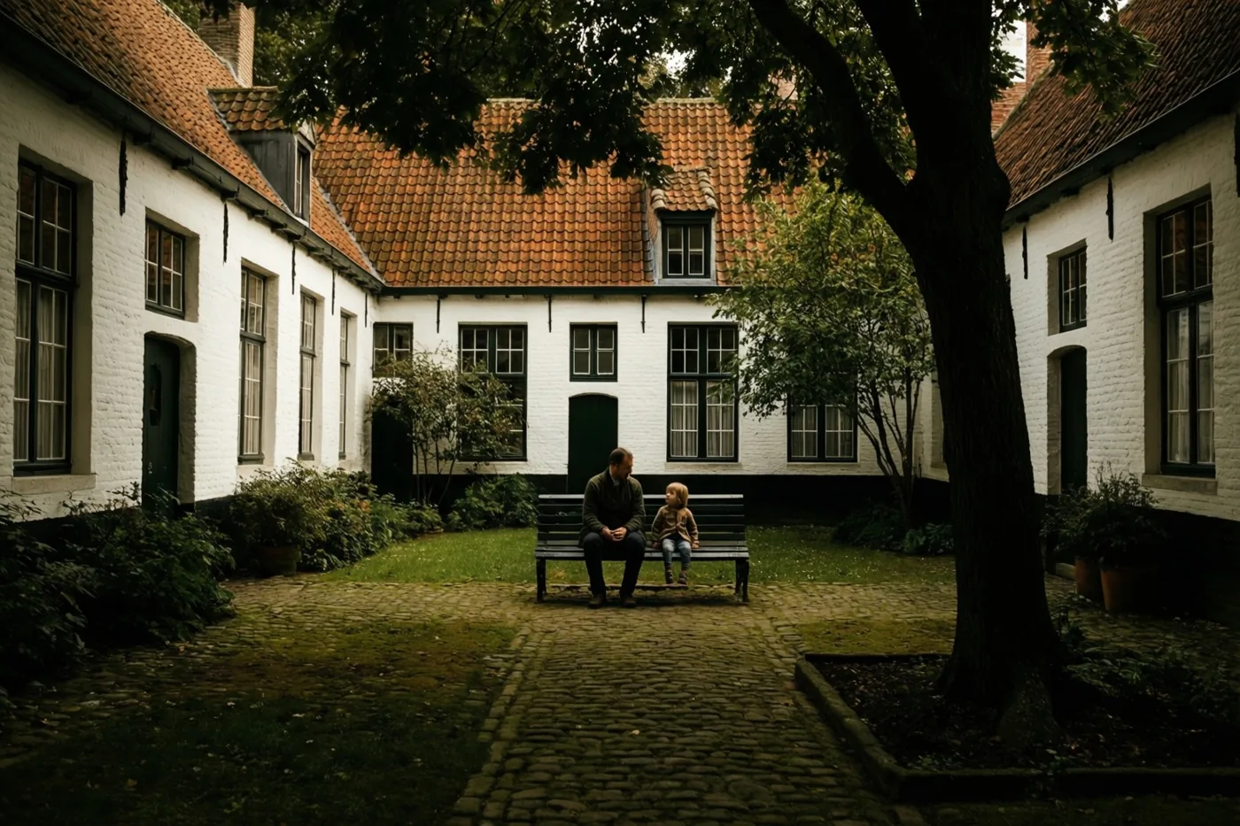 Een vader en kind zitten op een bank in een rustige binnenplaats met bomen, omringd door witte gebouwen met rode pannendaken.