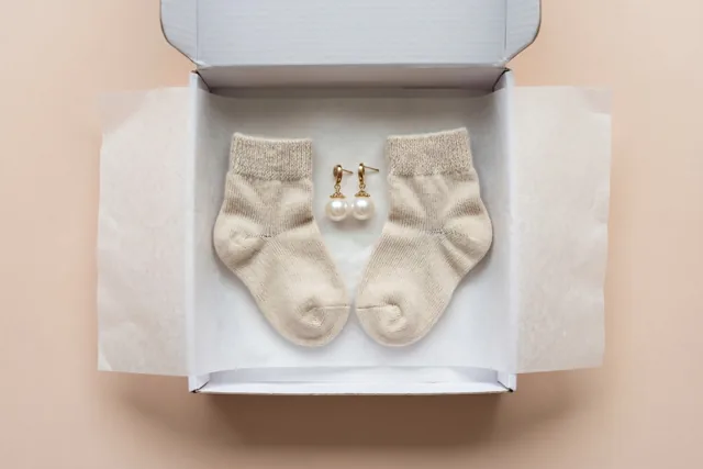 Een open witte geschenkdoos bevat beige babysokjes en pareloorbellen op wit zijdepapier tegen een zachte perzikkleurige achtergrond.