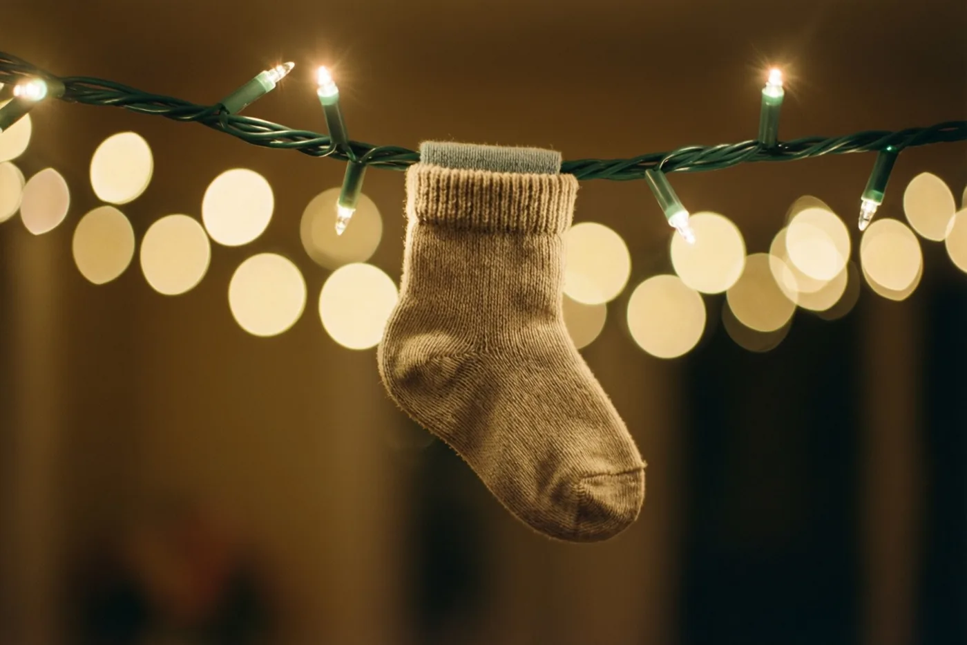Een beige babysokje hangt aan een snoer met groene lichtjes, tegen een wazige achtergrond met ronde lichtvlekken in groene en gouden tinten.