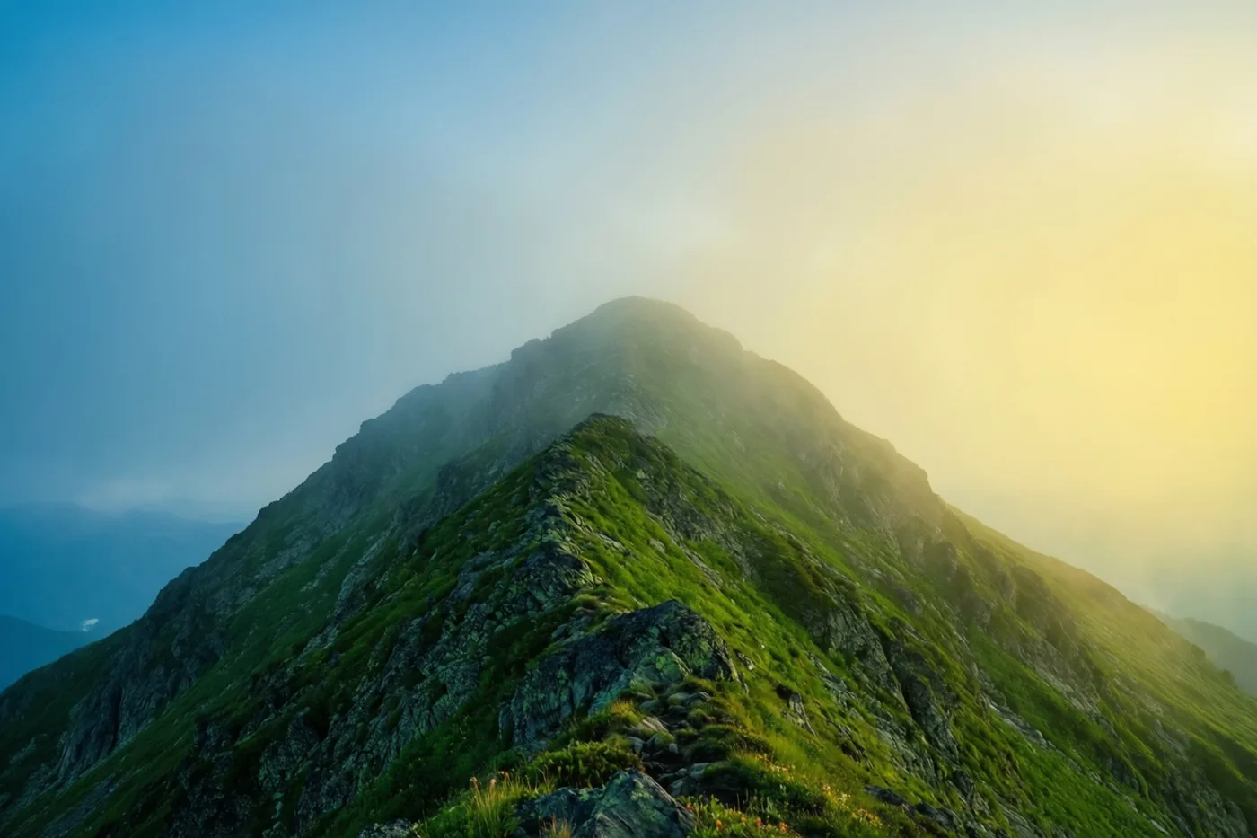 Een bergrug met groen gras en rotsen rijst op in zachte ochtendmist, verlicht door het warme gele licht van de opkomende zon.