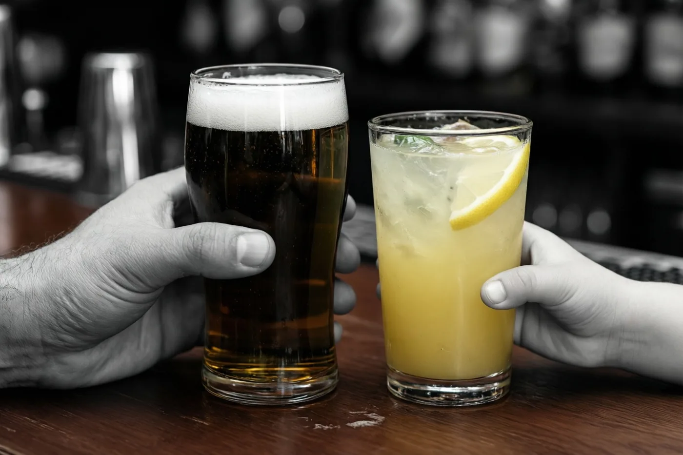 Een grote en een kleine hand houden donker bier en limonade met ijs en een schijfje citroen vast op een houten toog, met de dranken in kleur en de handen en achtergrond in zwart-wit.