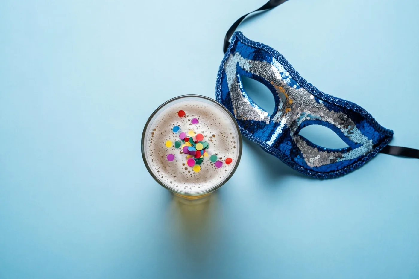 Van bovenaf is een glas bier met gekleurde confetti op het schuim te zien naast een blauw-zilveren paillettenmasker op een lichtblauwe ondergrond.