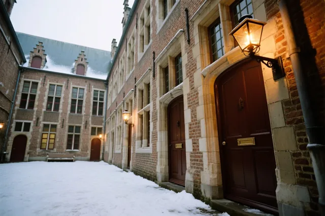 Op een besneeuwde binnenplaats van een historisch bakstenen gebouw staan houten boogdeuren met brandende lantaarns onder een bewolkte hemel.