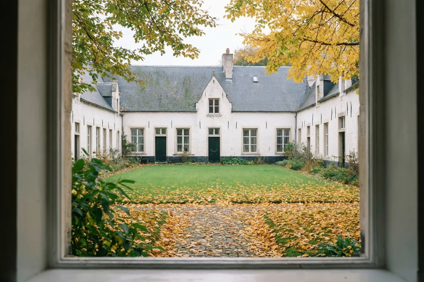 Door een raam is een binnenplein met witgekalkte huizen, groen gras en stenen paden vol gouden herfstbladeren te zien, omlijst door takken met vergelende bladeren.