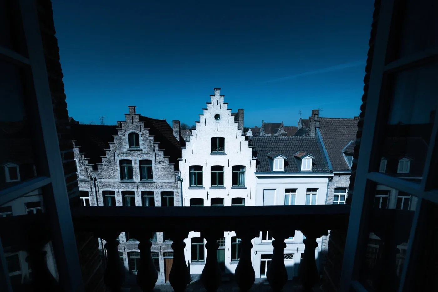 Vanaf een balkon zijn de daken van een oud Vlaams stadje te zien onder een heldere lucht, met een donkere balkonleuning op de voorgrond en overwegend diepblauwe tinten.