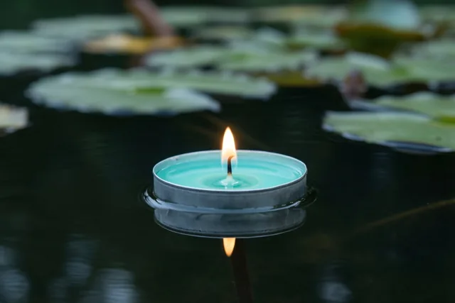 Een brandend theelichtje van blauwe was drijft op een donkere vijver tussen onscherpe groene waterleliebladeren, met een weerspiegeling van de vlam in het water.