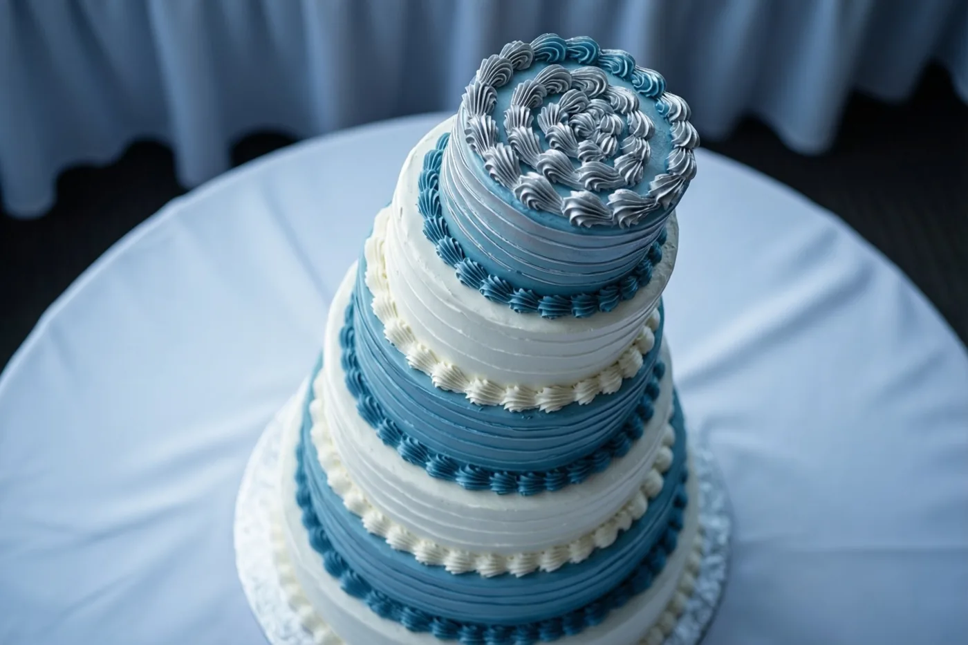 Een bruidstaart met meerdere lagen blauw en wit glazuur en fijn spuitwerk staat op een witte taartvoet op een wit tafelkleed, voor een onscherp blauw gordijn.