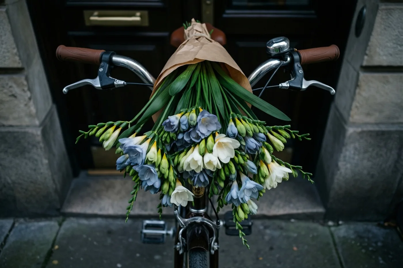 Een boeket blauwe en witte freesia's in kraftpapier hangt aan het stuur van een fiets met bruine leren handvatten voor een donkere deur.