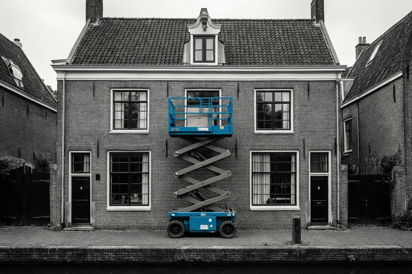 Een felblauwe schaarlift reikt tot een raam op de tweede verdieping van een bakstenen huis aan een kanaal, terwijl de rest van het beeld zwart-wit is.