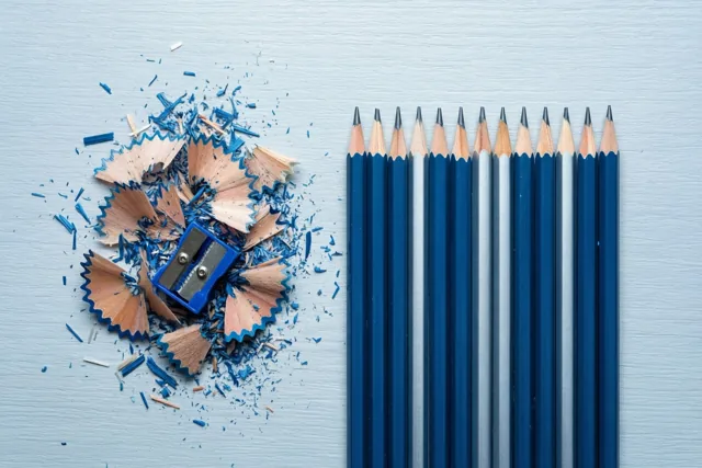 Een blauwe potloodslijper ligt tussen houtkleurig en blauw slijpsel naast een rij van vijftig geslepen potloden op een lichtblauwe ondergrond met textuur.