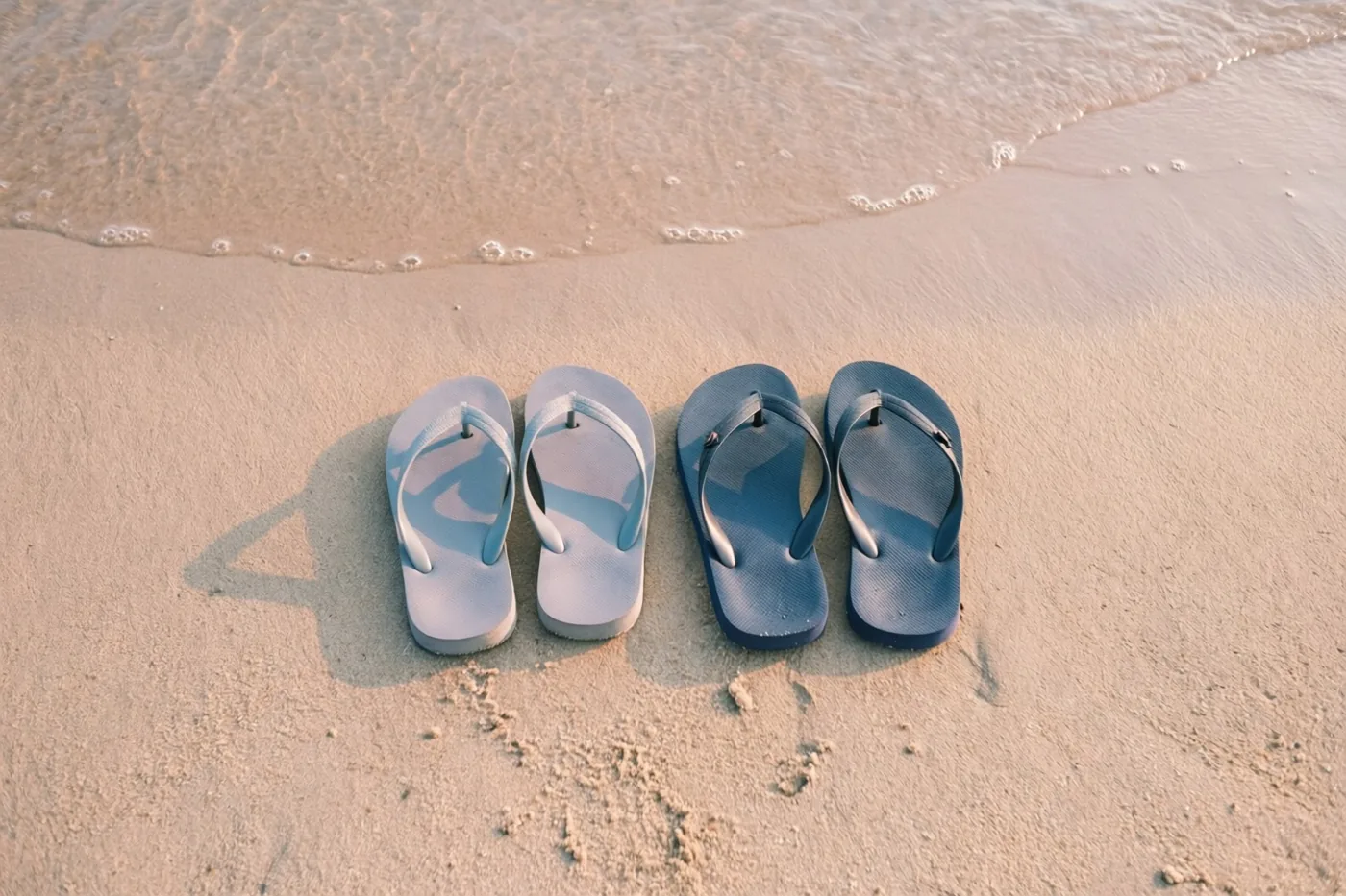 Een paar lichtblauwe en een paar donkerblauwe teenslippers liggen naast elkaar op nat zand aan de waterlijn in warm goudkleurig licht.