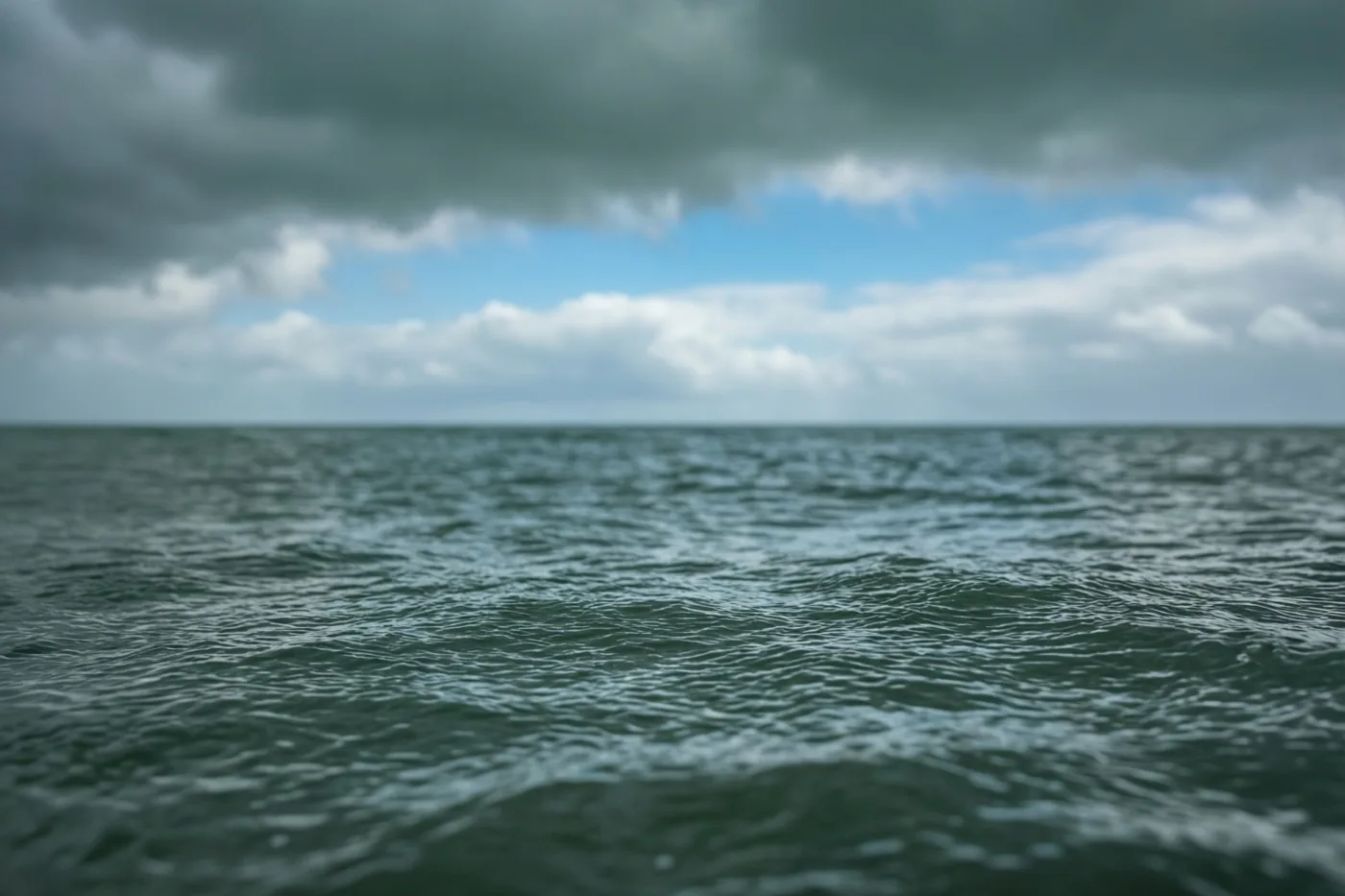 Donkere wolken boven een kalme zee wijken uiteen en laten een strook blauwe lucht zien in een blauwgrijs landschap.
