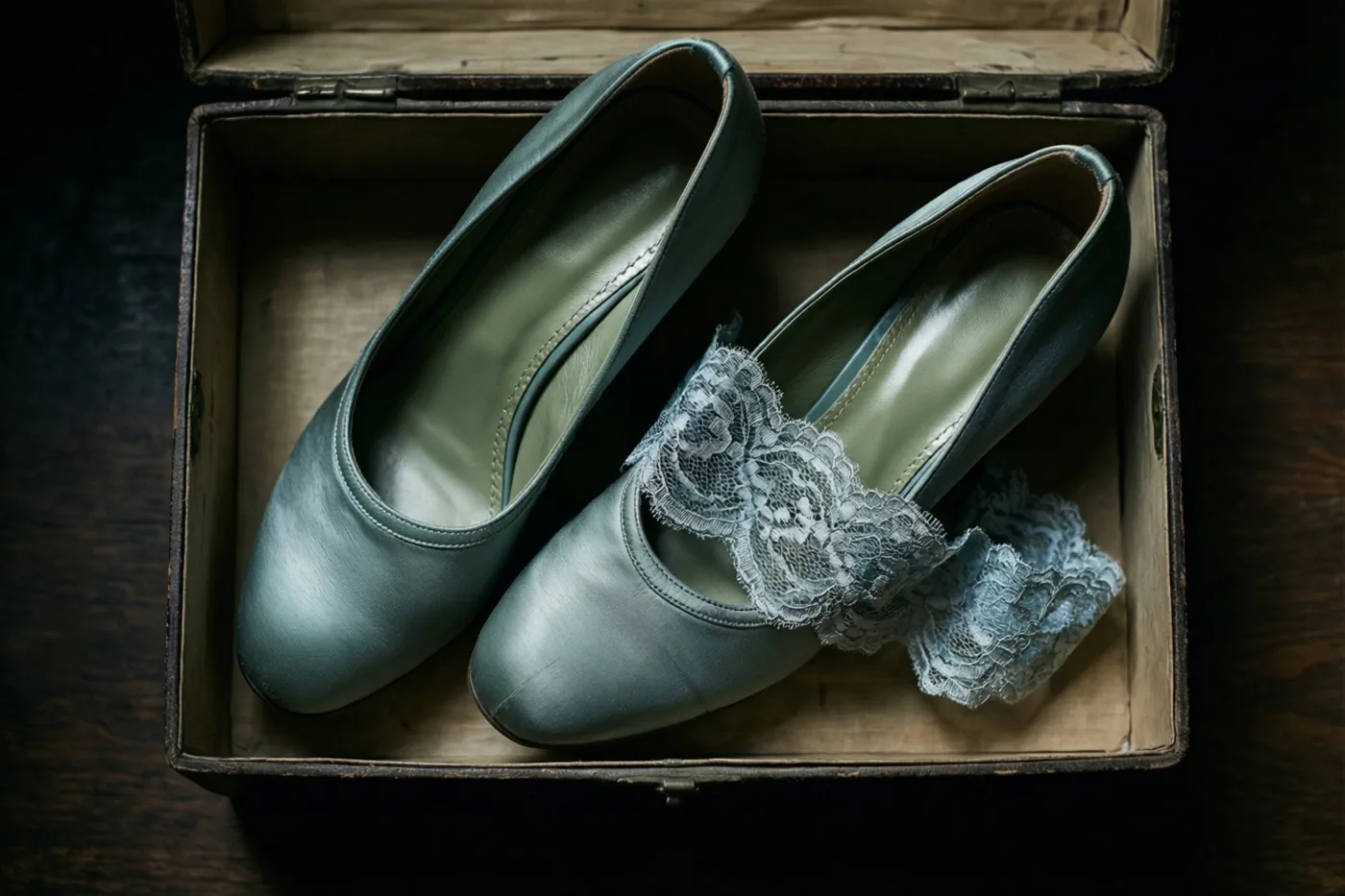 Blauwgroene satijnen bruidsschoenen in jarenvijftigstijl liggen met een kanten kousenband in een oude houten schoenendoos op een donker houten oppervlak.