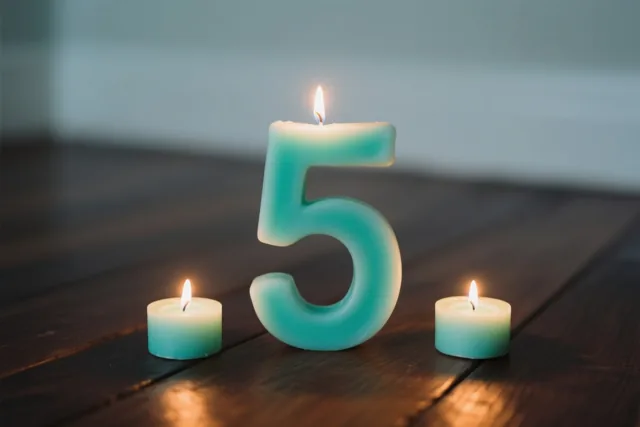 Een grote blauwgroene kaars in de vorm van een 5 en twee kleinere blauwgroene kaarsen branden op een donkere houten vloer tegen een donkere achtergrond.