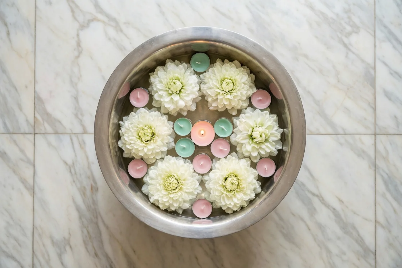 Een zilveren schaal met water, drijvende witte bloemen en kleurrijke theelichtjes staat op een witmarmeren vloer in zachte pasteltinten.