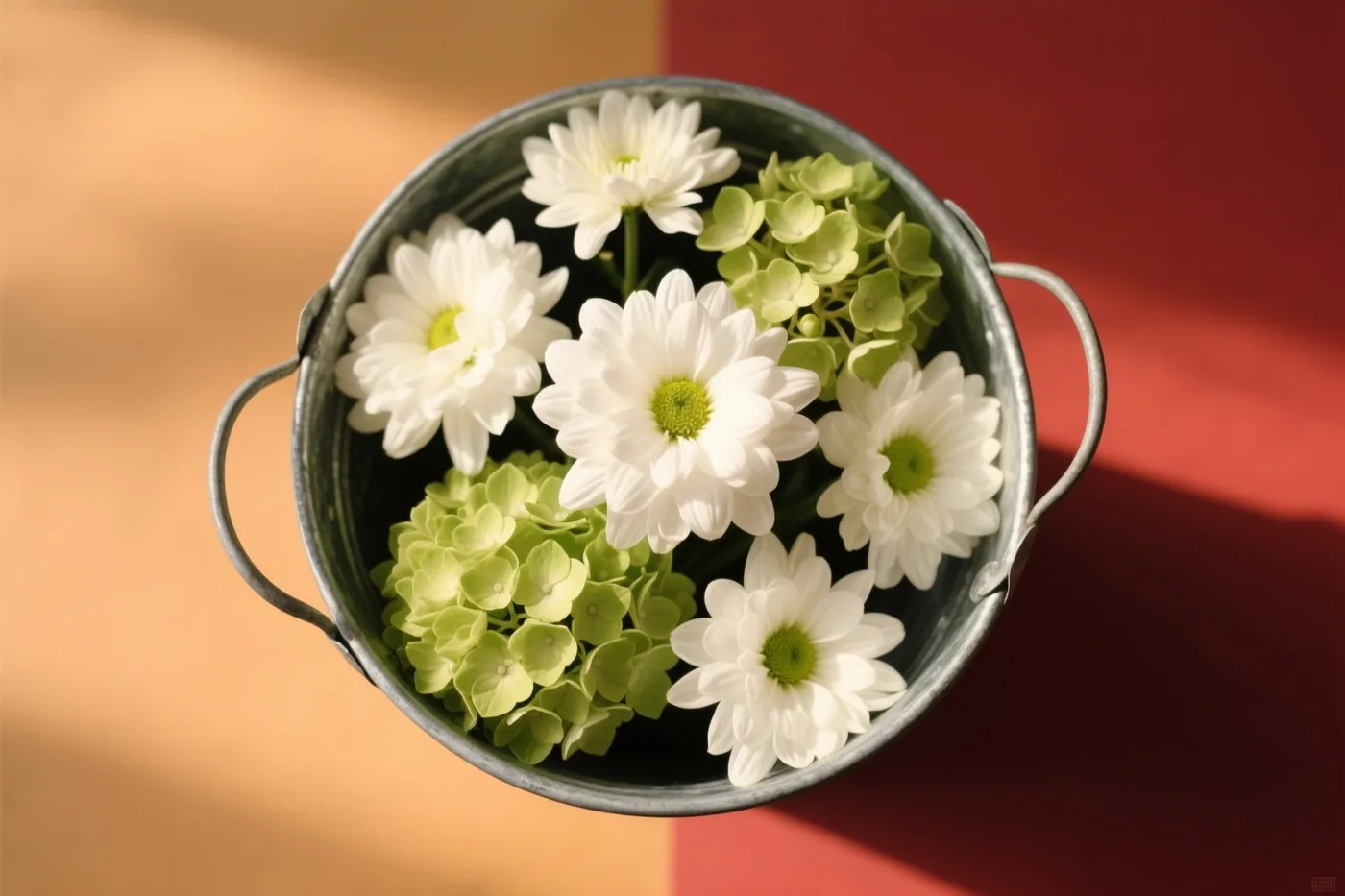 Van bovenaf zijn witte chrysanten en groene hortensiabloemen in een zinken emmer te zien tegen een achtergrond in beige en dieprood.