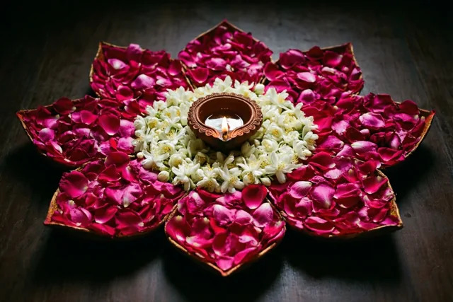 Roze rozenblaadjes en witte jasmijn vormen een lotusvormige rangoli rond een brandend olielampje op een donker houten oppervlak.