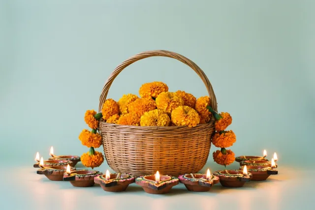 Een gevlochten mand vol feloranje bloemen staat in een kring van brandende lampjes van klei op een lichtblauwe ondergrond voor een lichtblauwe achtergrond.