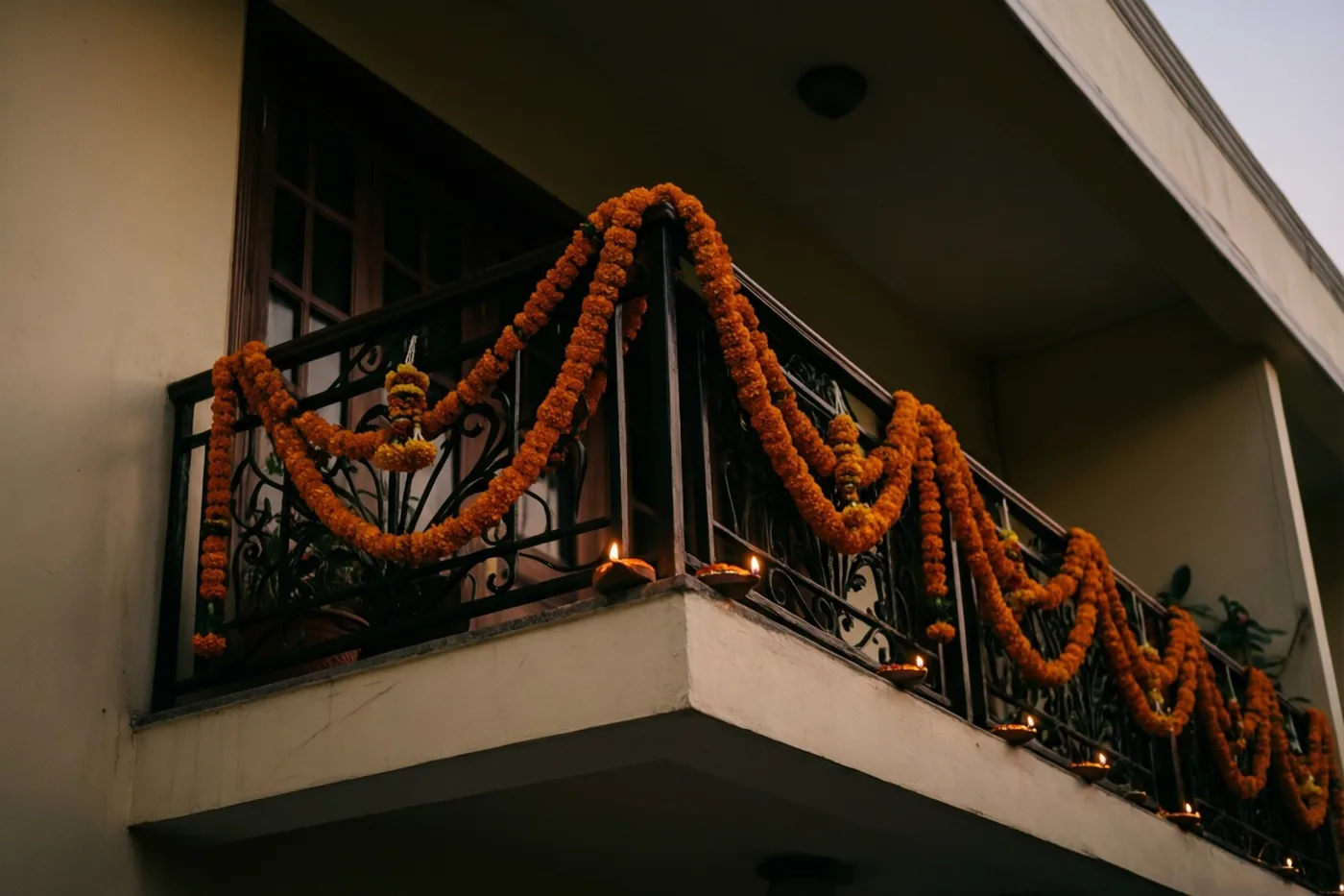 Slingers van oranje afrikaantjes hangen langs een balkonleuning met kleine brandende olielampjes, voor een gevel bij schemering.