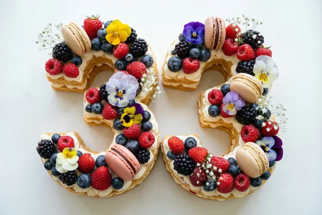 Een taart in de vorm van 33 staat op een effen wit oppervlak en is versierd met bessen, macarons en eetbare bloemen.