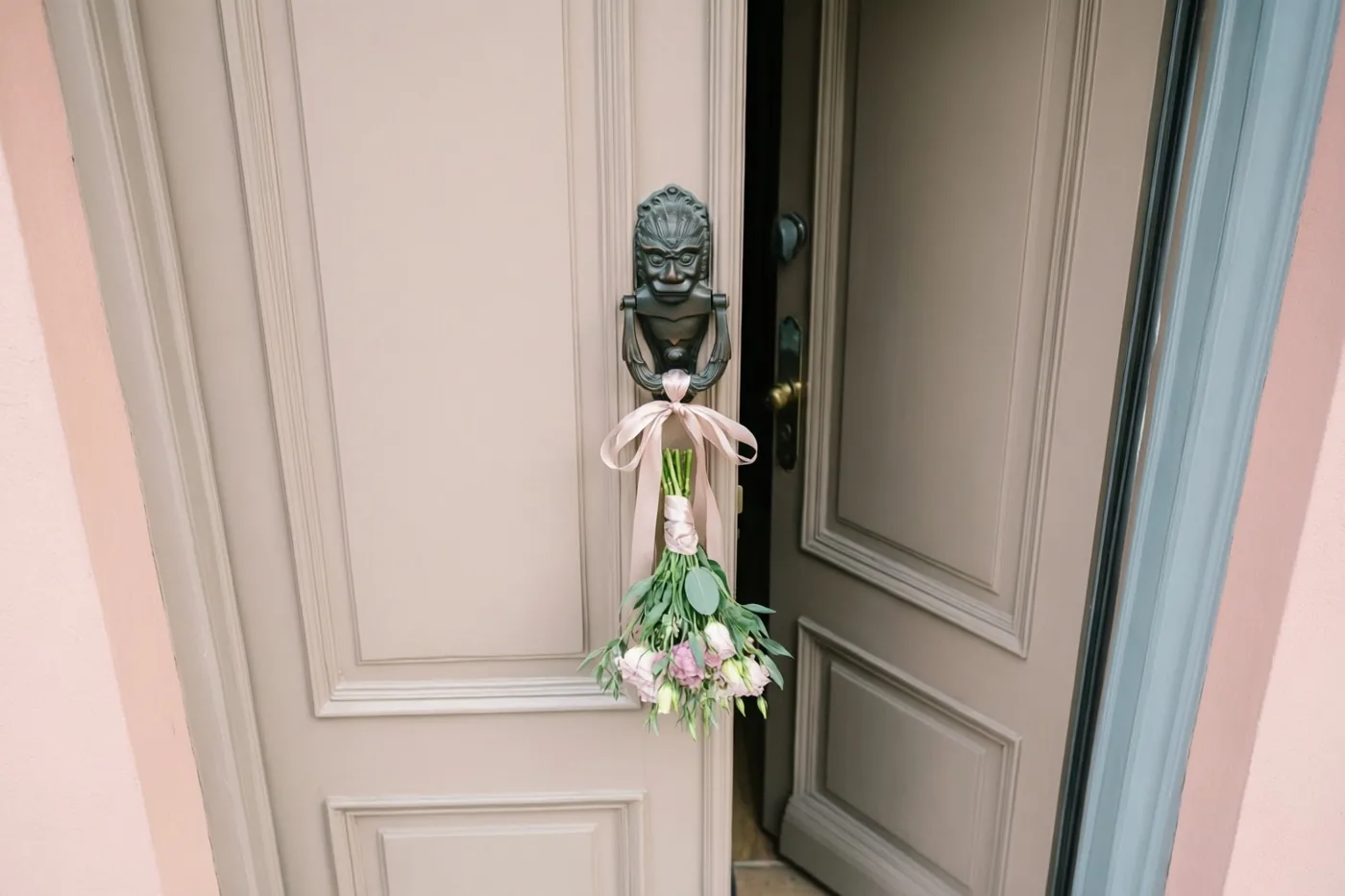 Een boeketje roze en witte bloemen met een lichtroze lint hangt aan een zwarte deurklopper op een lichtbeige voordeur die op een kier staat, met roze en blauwgrijze muren eromheen.